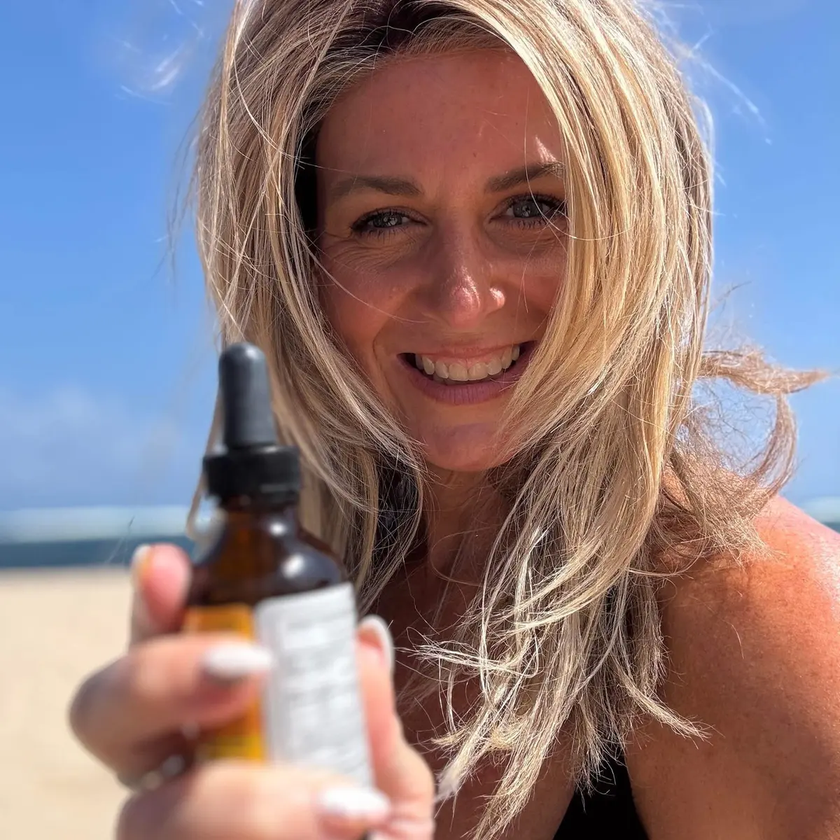 Dr. Kristal smiling on the beach, holding a bottle of the Drops toward the camera